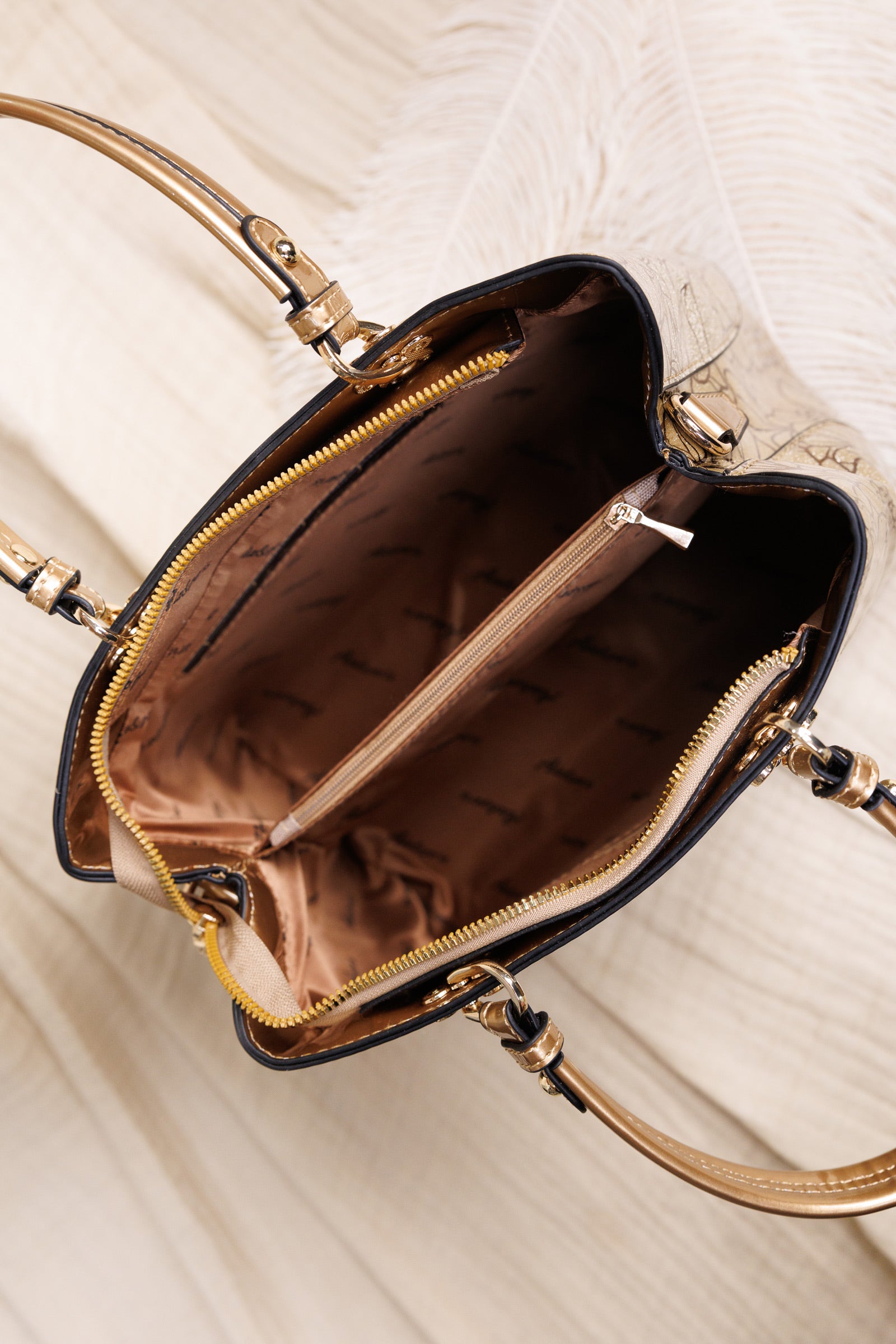 An overhead close-up of a structured handbag interior showing a black exterior, gold-tone hardware, and a brown satin-lined compartment with a zip pocket.