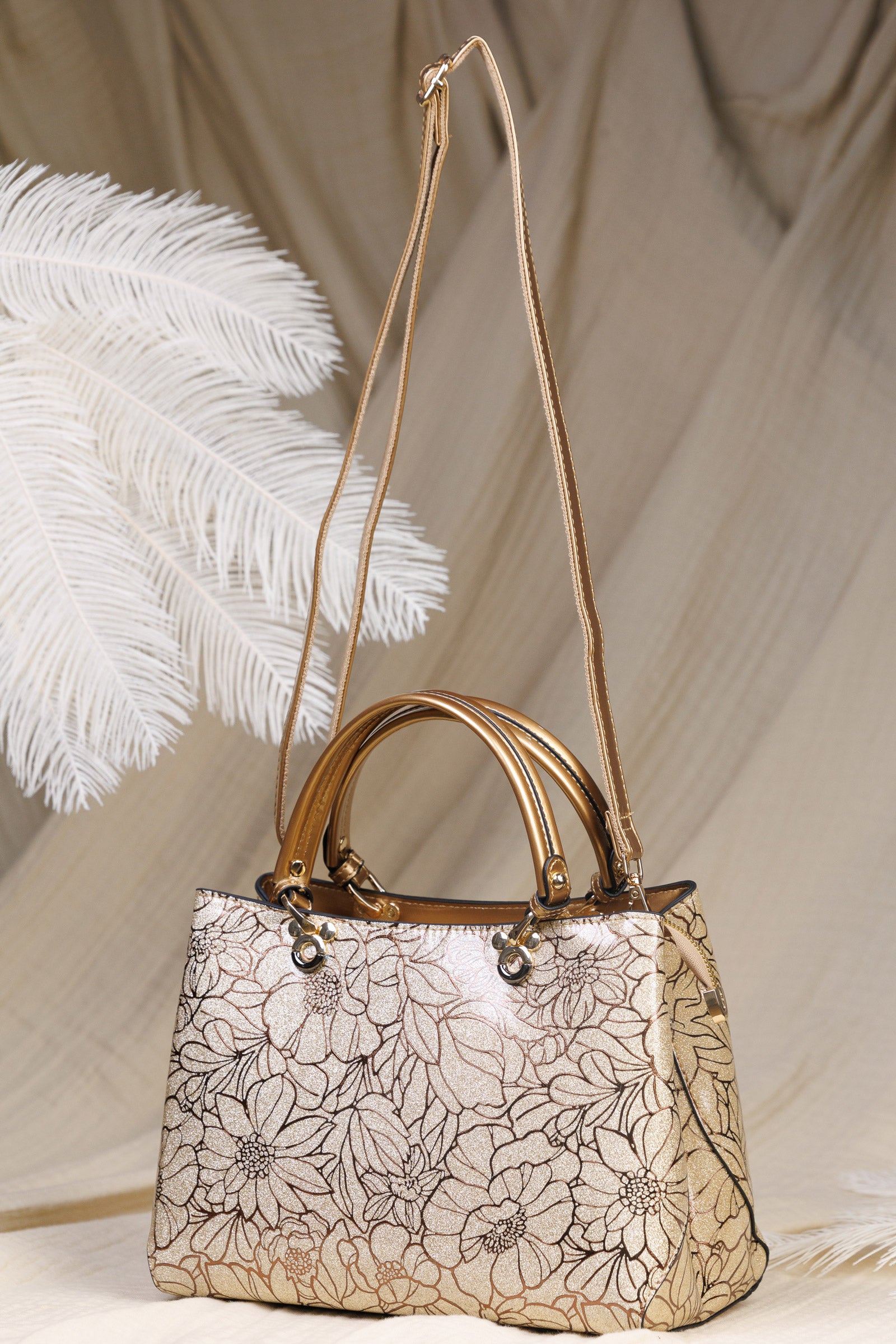 A structured handbag with a metallic gold floral pattern on a beige base, featuring dual rolled top handles and a long shoulder strap, styled with white decorative feathers against a draped beige fabric backdrop.