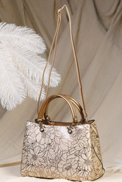 A structured handbag with a metallic gold floral pattern on a beige base, featuring dual rolled top handles and a long shoulder strap, styled with white decorative feathers against a draped beige fabric backdrop.