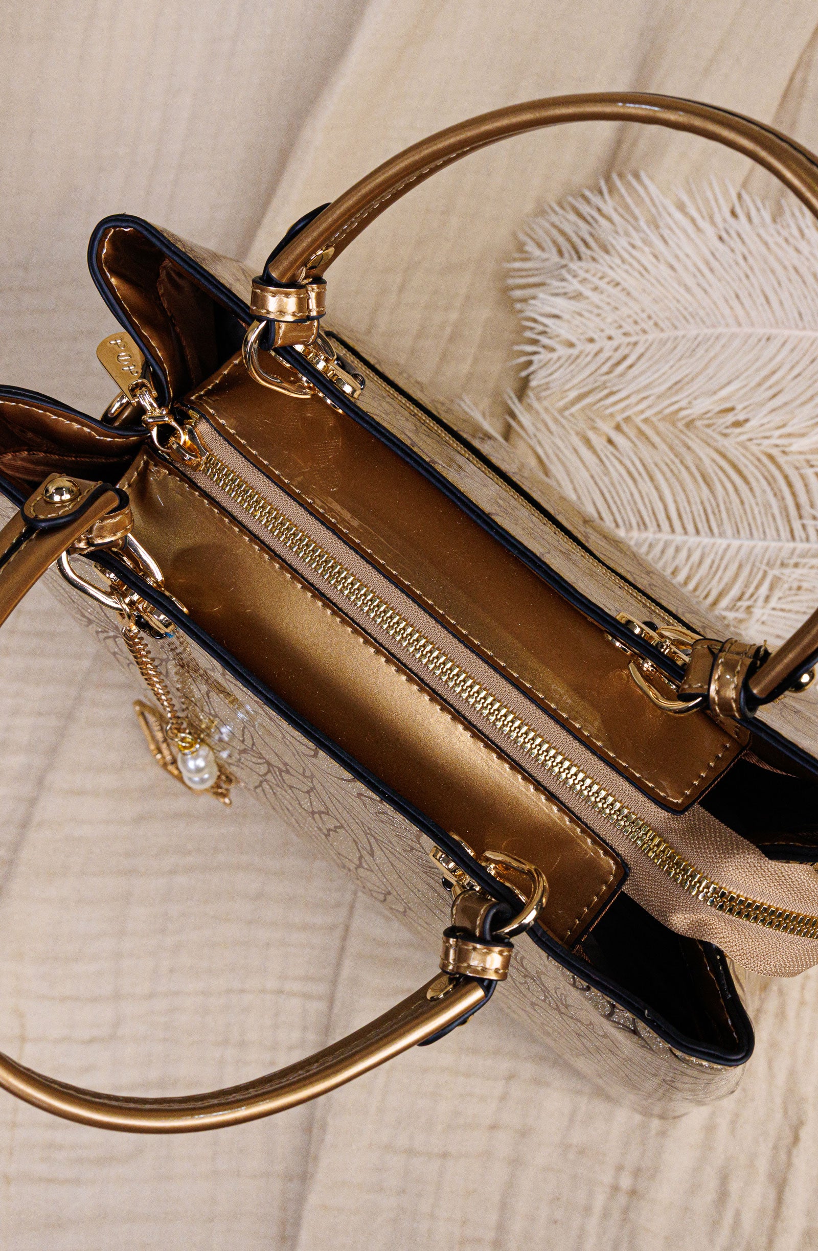 A close-up of a structured handbag with metallic gold-tone hardware, a gold zipper, and a beige textured body, styled on a light fabric surface with a white feather prop.