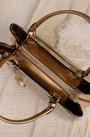 A close-up of a structured handbag with metallic gold-tone hardware, a gold zipper, and a beige textured body, styled on a light fabric surface with a white feather prop.