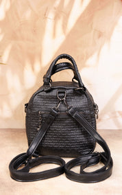 A black textured faux-leather mini backpack with top handles, a front zip pocket, and a detachable shoulder strap, displayed against a warm beige background with leaf shadows.