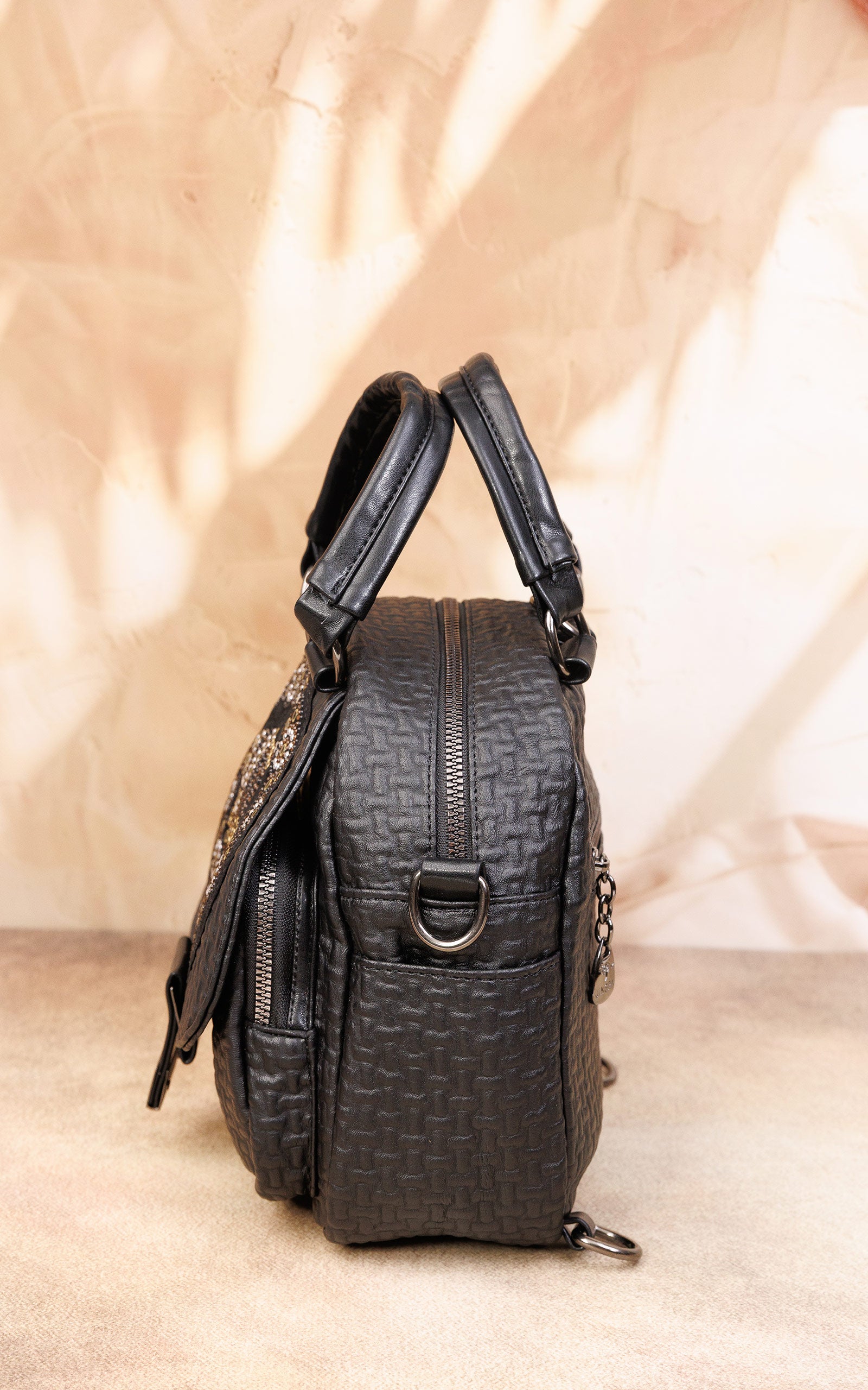 A black textured leather handbag with top handles and a front zipper pocket, photographed against a warm beige backdrop with soft leaf shadows.