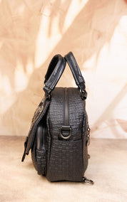 A black textured leather handbag with top handles and a front zipper pocket, photographed against a warm beige backdrop with soft leaf shadows.
