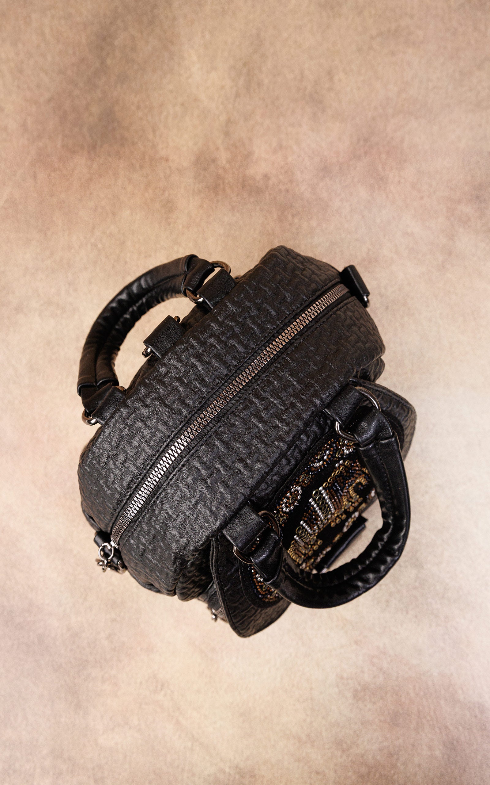 A black quilted leather handbag with a top zipper and double top handles, photographed from a top-down angle on a textured beige background.
