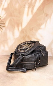 A black textured faux-croc shoulder bag with a large oval rhinestone-embellished flap, resting on a beige surface with warm leaf shadows cast across it.