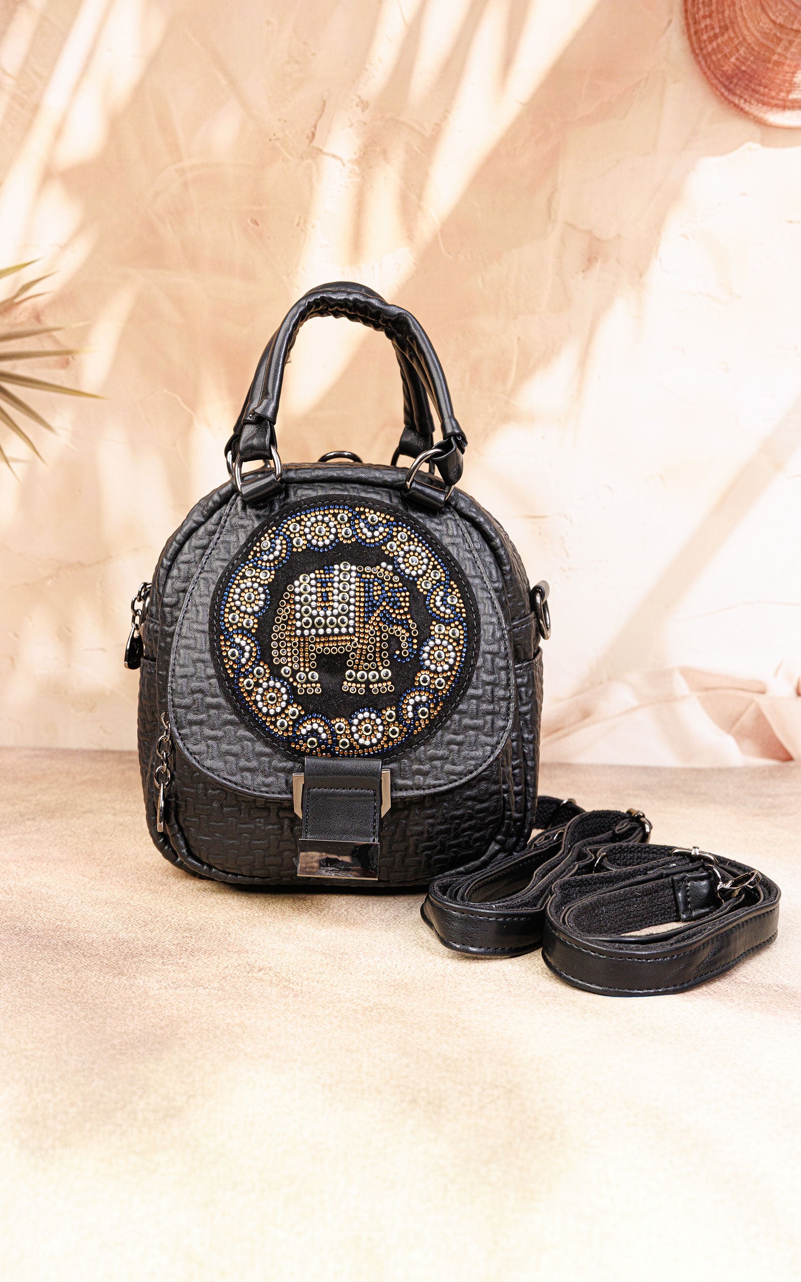A black textured leather handbag with a circular crystal-embellished front logo, dual top handles, and a detachable shoulder strap, displayed on a beige surface with warm shadow overlays.