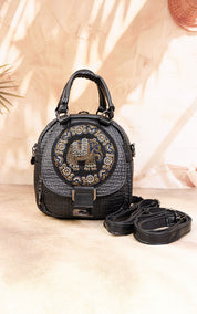 A black textured leather handbag with a circular crystal-embellished front logo, dual top handles, and a detachable shoulder strap, displayed on a beige surface with warm shadow overlays.