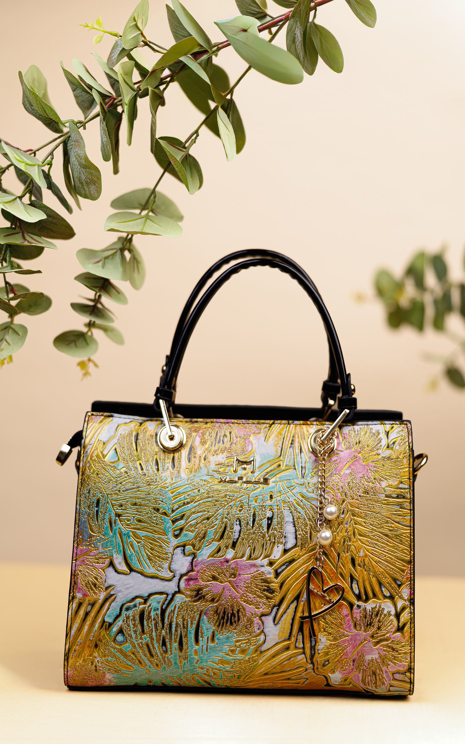 A structured handbag with a metallic gold foil palm leaf pattern, pink floral accents, turquoise details, and black leather top handles, styled with eucalyptus branches against a beige background.