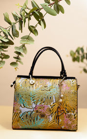 A structured handbag with a metallic gold foil palm leaf pattern, pink floral accents, turquoise details, and black leather top handles, styled with eucalyptus branches against a beige background.