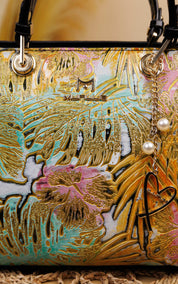 A close-up of a designer handbag featuring a highly textured, multicolor embossed pattern with gold leaf-like detailing, pink floral elements, and turquoise accents. The bag has black leather handles and a dangling charm with pearl beads and a black heart outline.