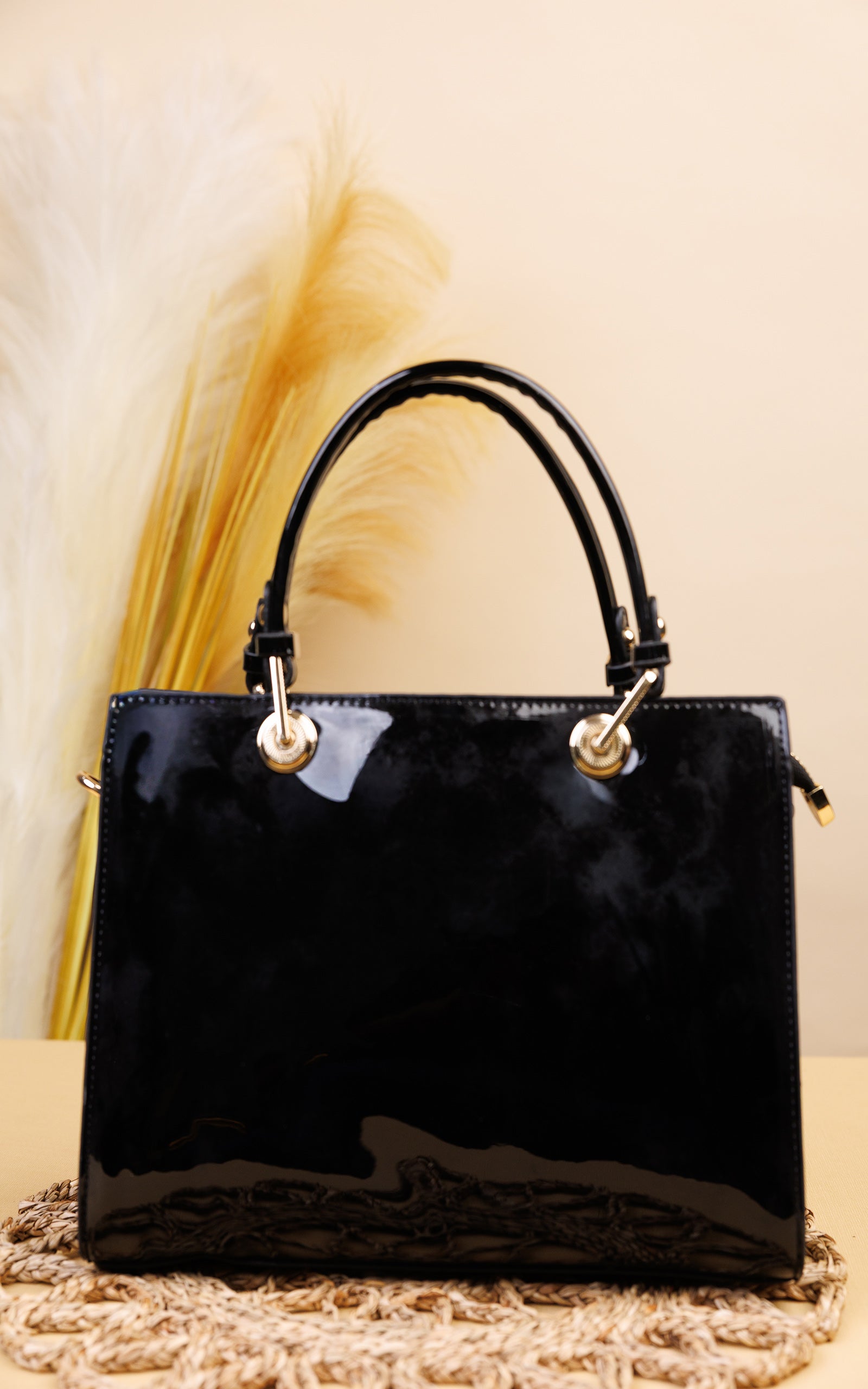 A glossy black patent leather handbag with double top handles and gold-tone hardware, displayed on a woven mat against a beige background with dried pampas grass.