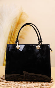 A glossy black patent leather handbag with double top handles and gold-tone hardware, displayed on a woven mat against a beige background with dried pampas grass.