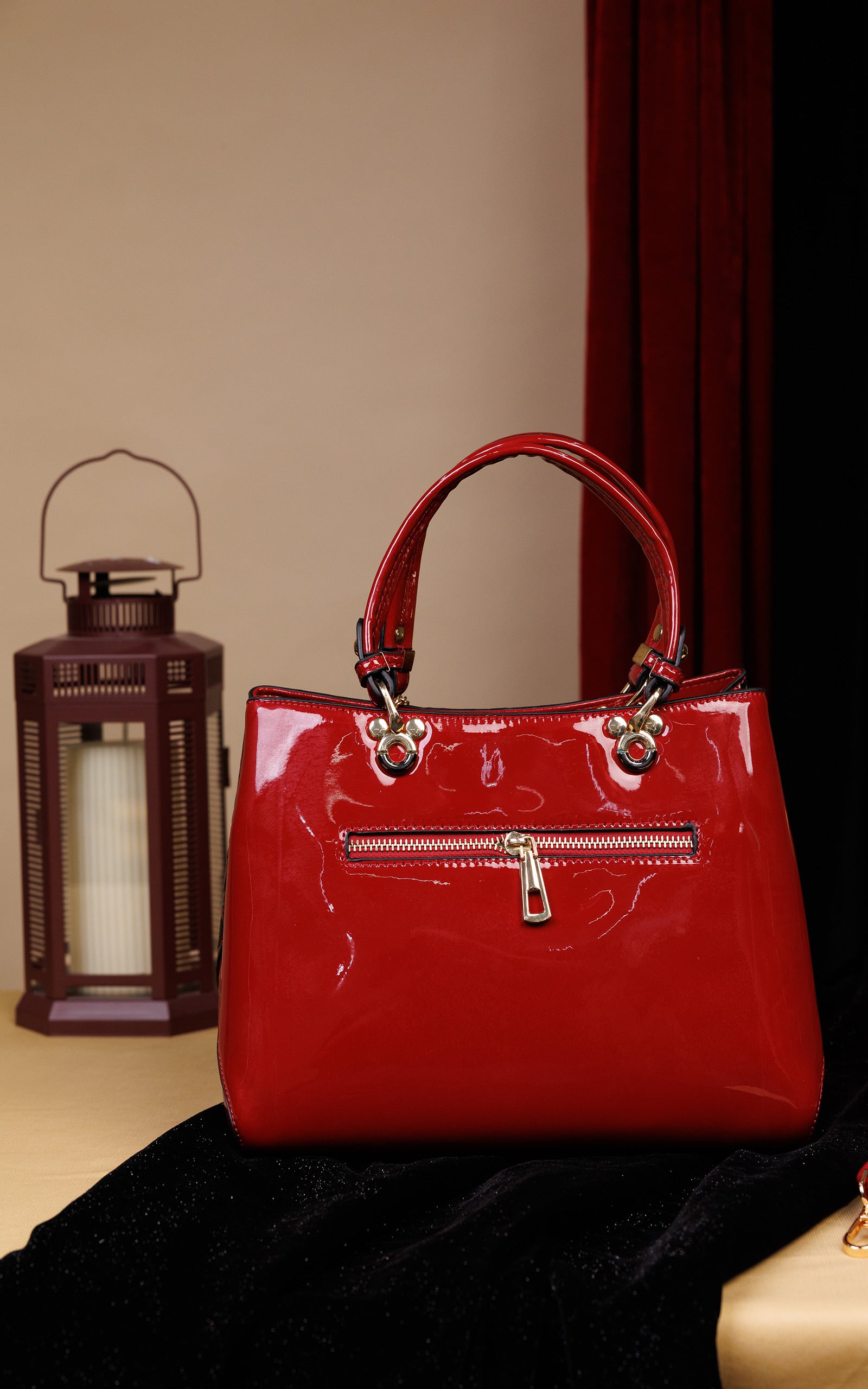 A glossy red patent leather handbag with double top handles and gold-tone hardware, displayed on a black velvet surface next to a lantern against a beige and dark red background.