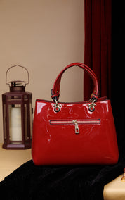 A glossy red patent leather handbag with double top handles and gold-tone hardware, displayed on a black velvet surface next to a lantern against a beige and dark red background.