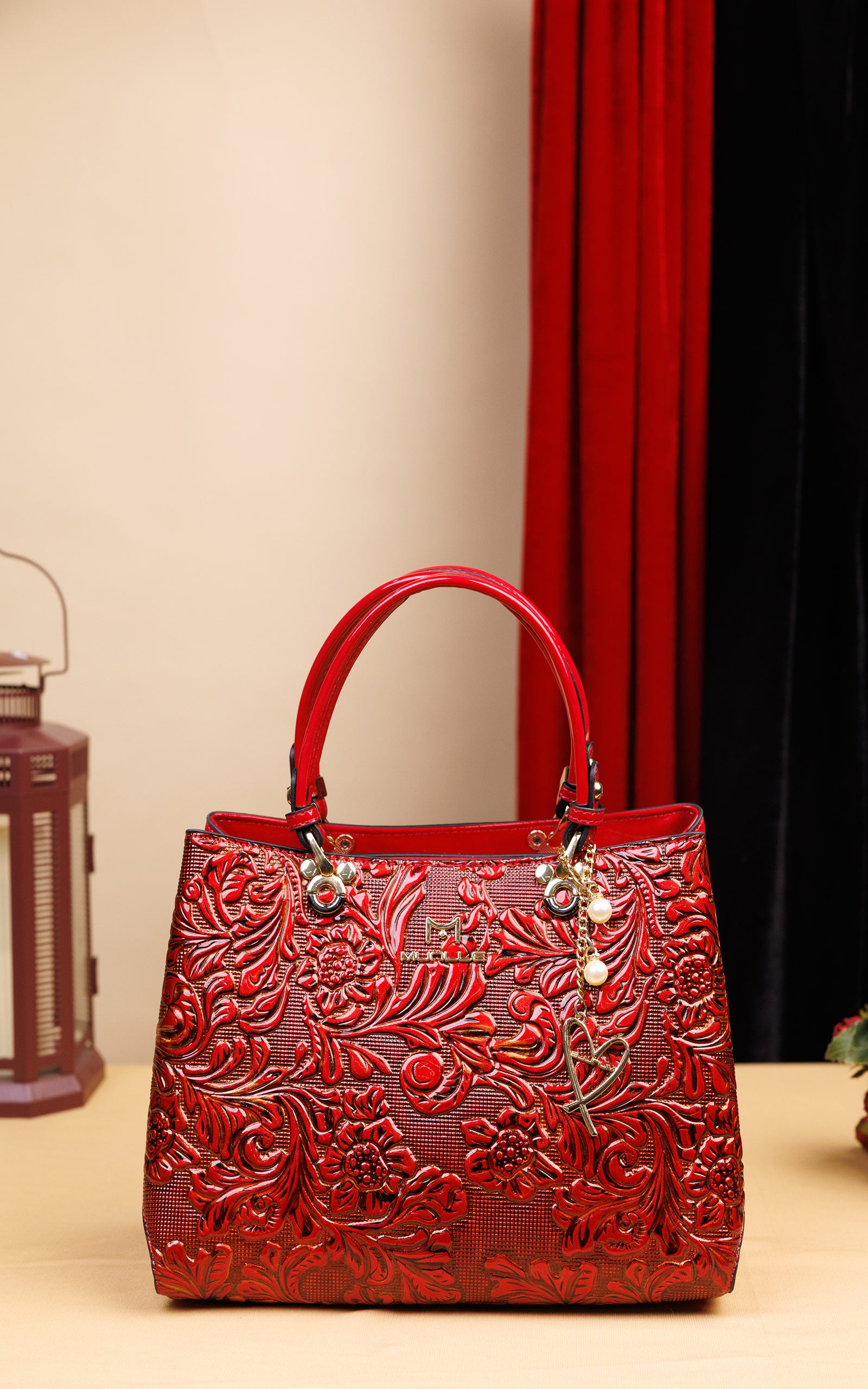 A red embossed leather handbag with floral patterns, dual top handles, and a hanging charm, displayed on a tabletop with a beige wall and red curtain in the background.