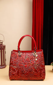 A red embossed leather handbag with floral patterns, dual top handles, and a hanging charm, displayed on a tabletop with a beige wall and red curtain in the background.