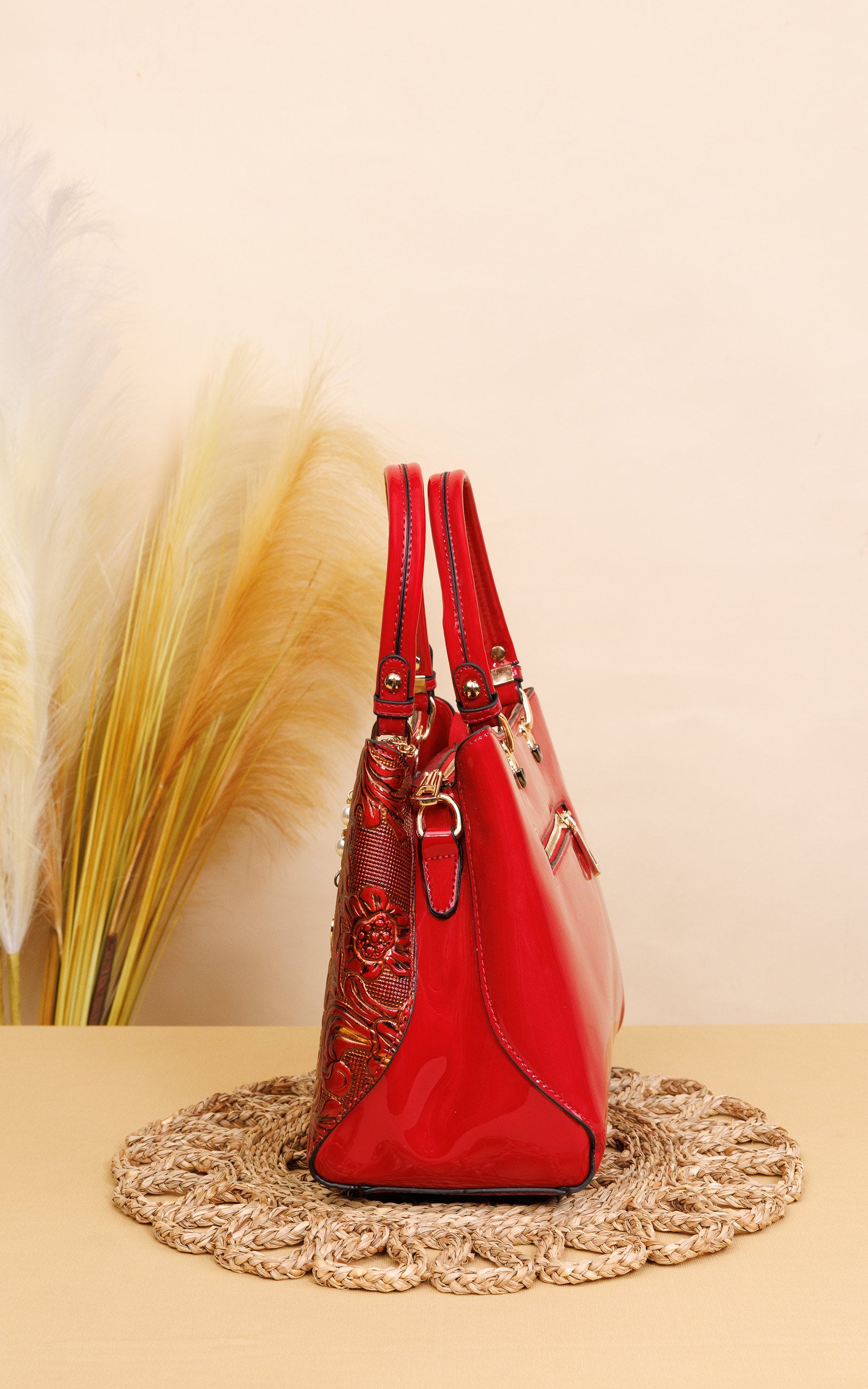 A red patent leather handbag with gold-tone hardware and a textured red patterned side panel, displayed on a woven jute mat against a beige background with dried pampas grass.