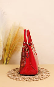 A red patent leather handbag with gold-tone hardware and a textured red patterned side panel, displayed on a woven jute mat against a beige background with dried pampas grass.