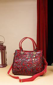 A red embossed leather handbag with floral detailing, dual top handles, and a matching red shoulder strap, displayed on a tabletop against a beige wall with a red curtain in the background.