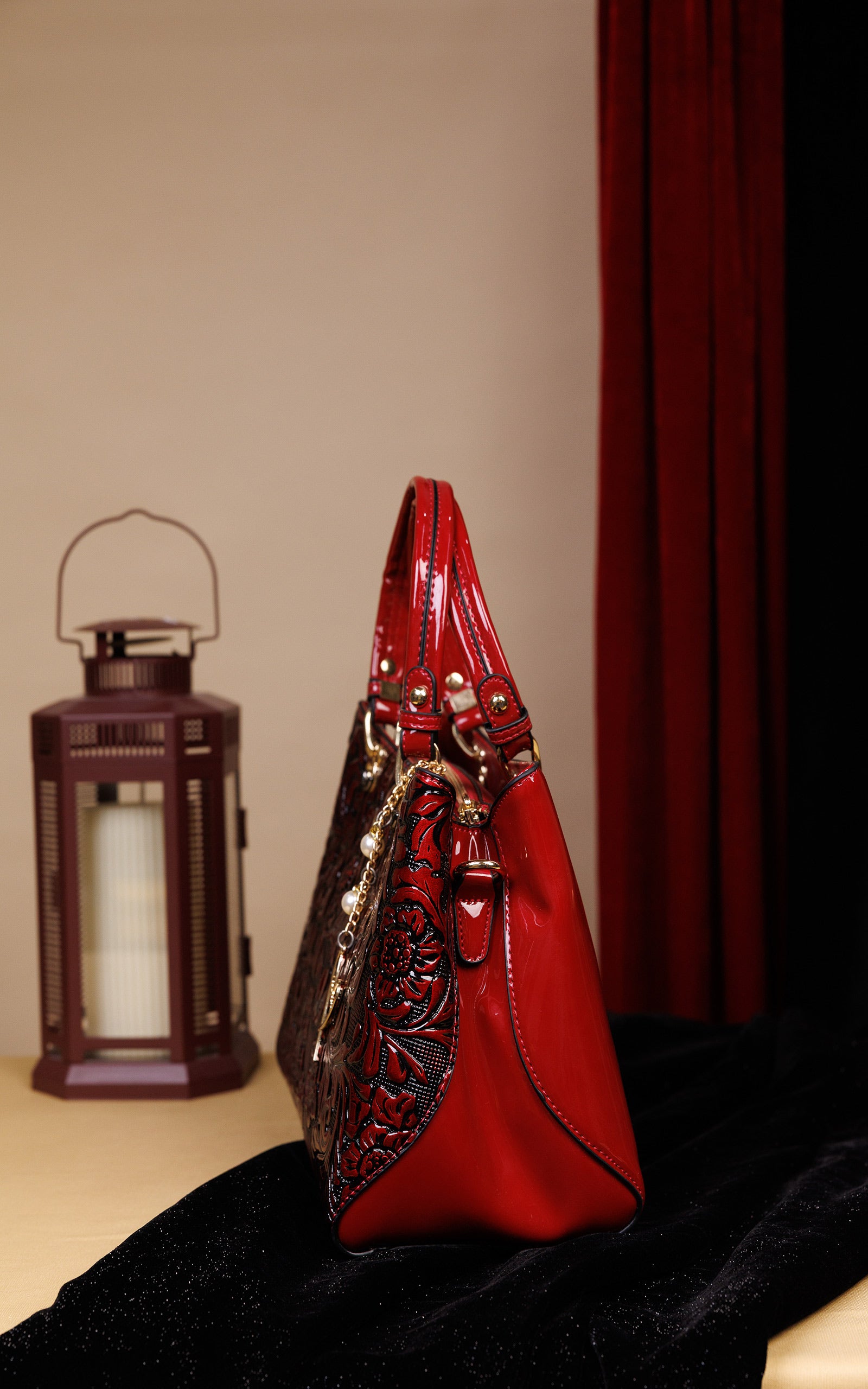 A red leather handbag with black floral embroidery and gold-tone hardware, standing on a black velvet surface next to a lit lantern against a beige wall and dark red curtain.