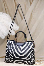 A structured handbag with a black and white zebra print body, black leather top handles and shoulder strap, and gold-tone hardware, styled on a beige draped fabric background with decorative white feathers and pearls.