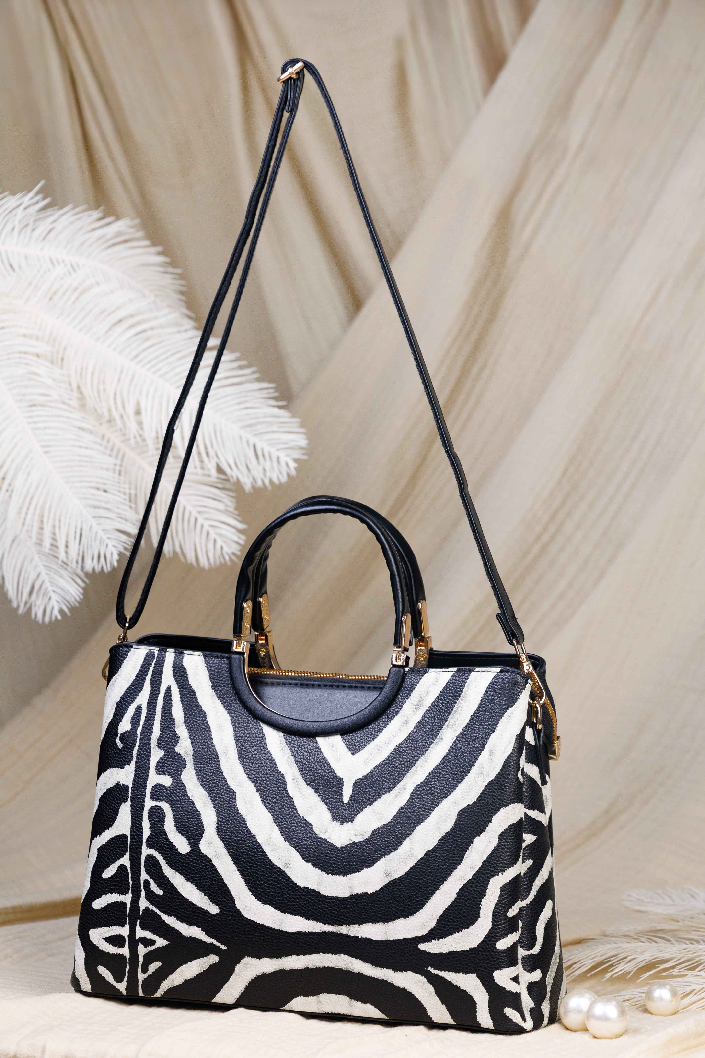 A structured handbag with a black and white zebra print body, black leather top handles and shoulder strap, and gold-tone hardware, styled on a beige draped fabric background with decorative white feathers and pearls.