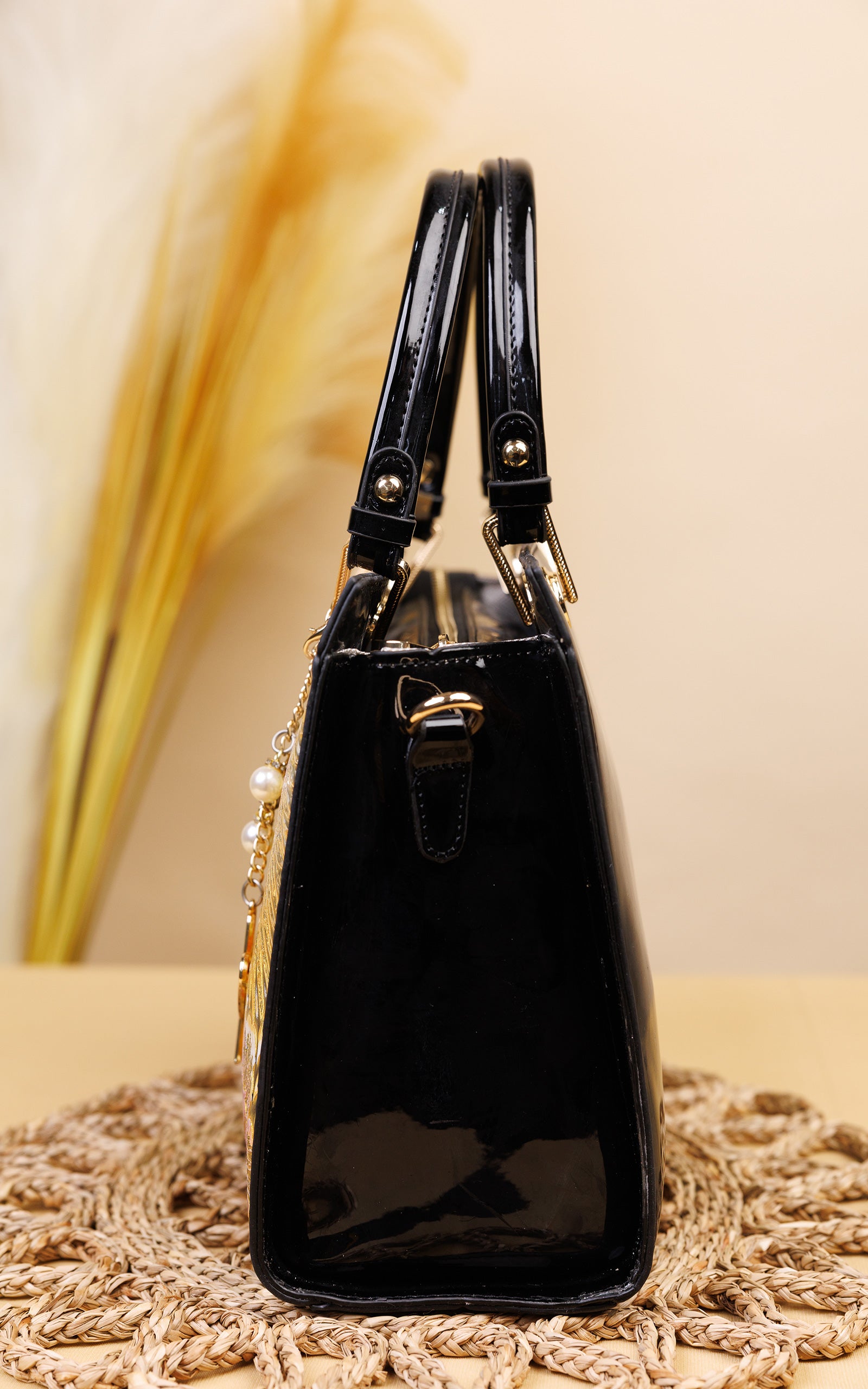 A glossy black patent leather handbag with gold-tone hardware and pearl accents, displayed on a woven beige mat against a warm beige background with dried grass.