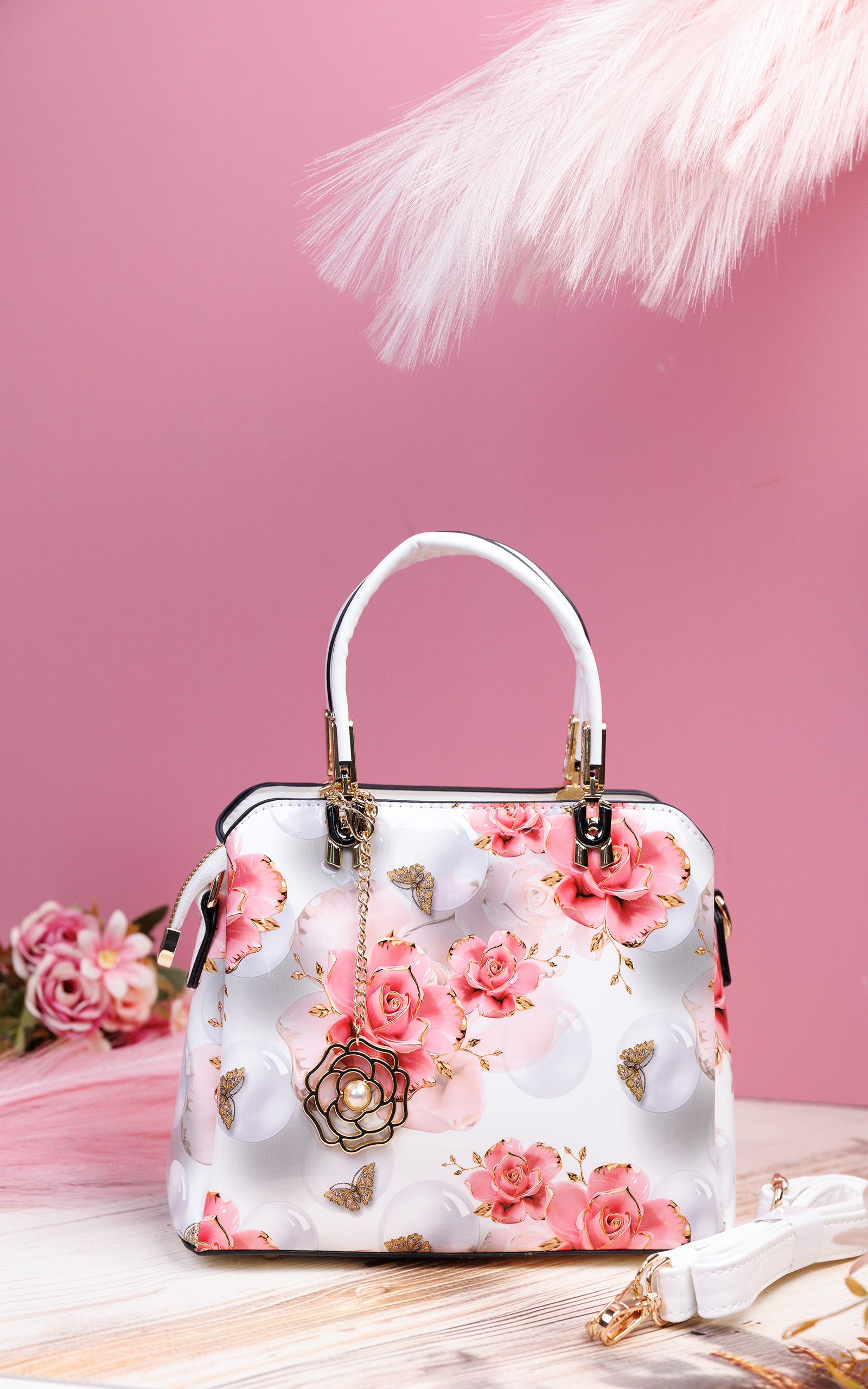 A structured white handbag with a pink rose floral print, featuring a white top handle, gold-tone hardware, and a decorative hanging flower charm, set against a pink background with soft decorative props.
