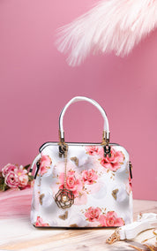 A structured white handbag with a pink rose floral print, featuring a white top handle, gold-tone hardware, and a decorative hanging flower charm, set against a pink background with soft decorative props.