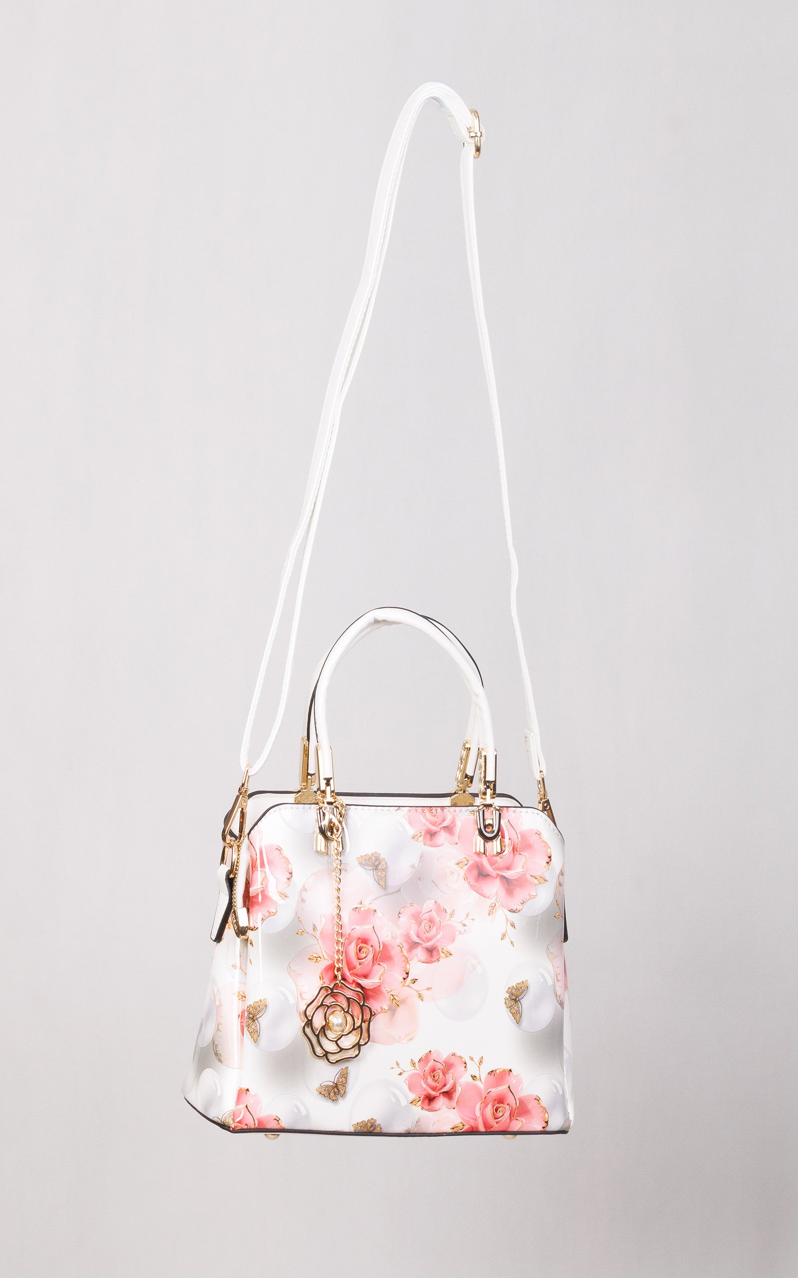 A structured white handbag with a pink floral print, featuring a long white shoulder strap, top handles, gold-tone hardware, and a hanging rose-shaped charm, shown against a light gray background.