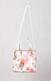 A structured white handbag with a pink floral print, featuring a long white shoulder strap, top handles, gold-tone hardware, and a hanging rose-shaped charm, shown against a light gray background.