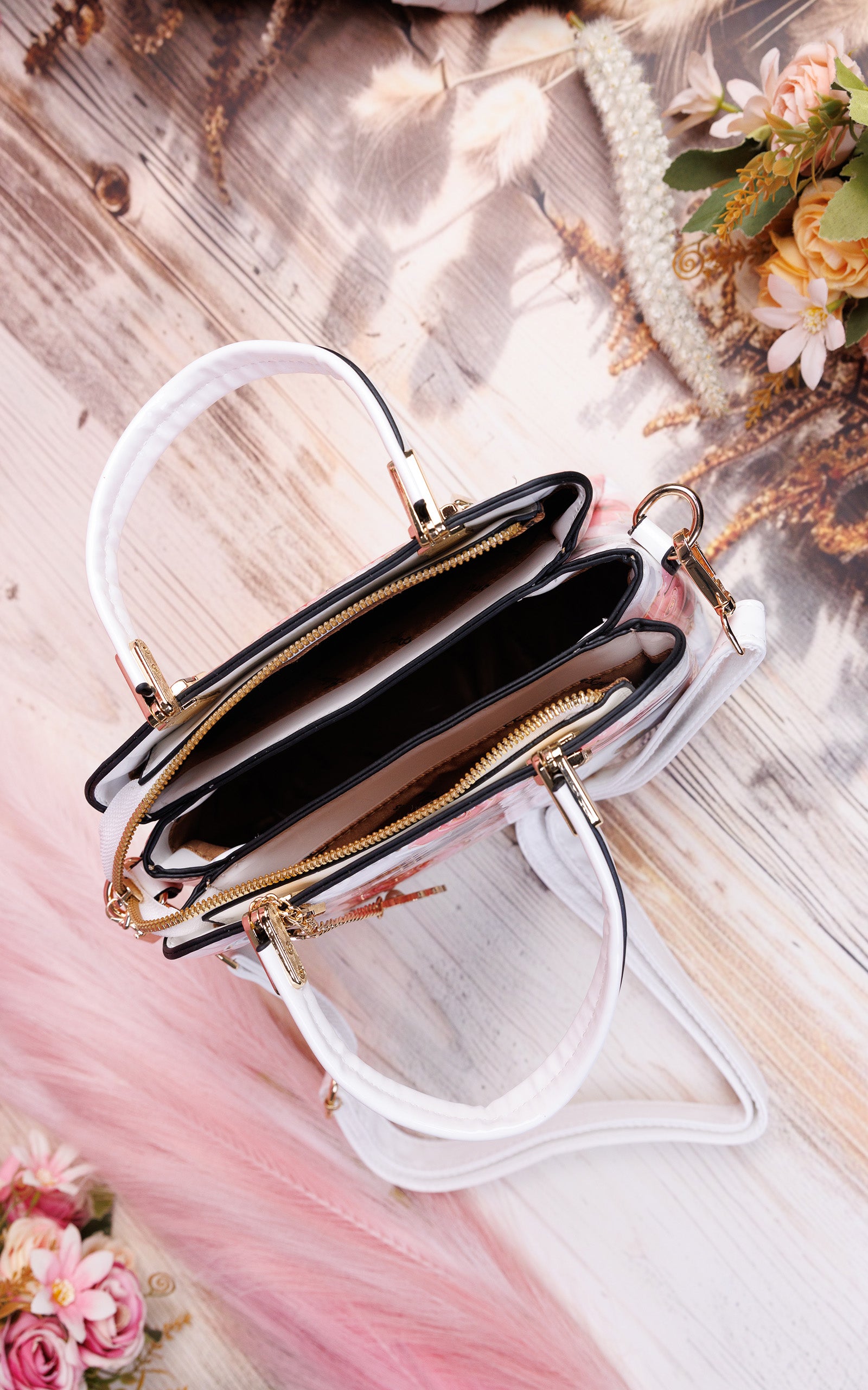 A clear vinyl handbag with white handles and gold-tone hardware, shown open on a styled wooden surface with pink fabric and dried floral accents.