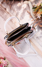 A clear vinyl handbag with white handles and gold-tone hardware, shown open on a styled wooden surface with pink fabric and dried floral accents.