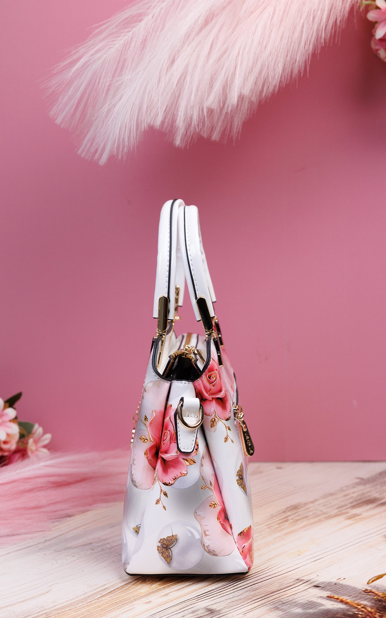 A structured handbag with a white base and a pink rose floral print, featuring gold-tone hardware and dual top handles, set against a pink background with decorative pampas grass.