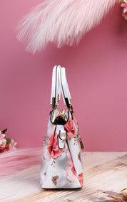 A structured handbag with a white base and a pink rose floral print, featuring gold-tone hardware and dual top handles, set against a pink background with decorative pampas grass.