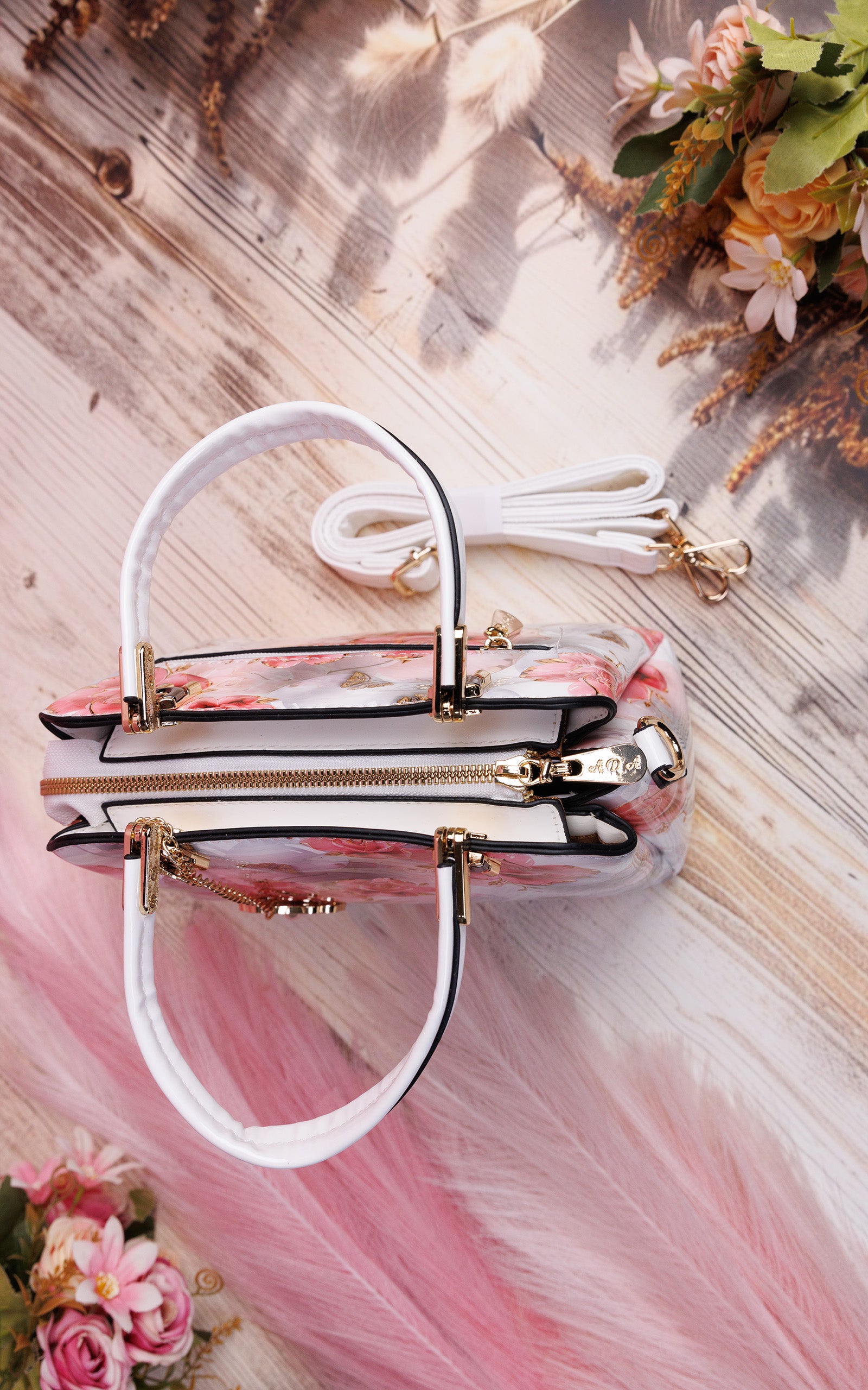 A clear acrylic handbag with a pink floral print, white handles, and gold-tone hardware, styled on a rustic wooden background with pink feathers and flowers.