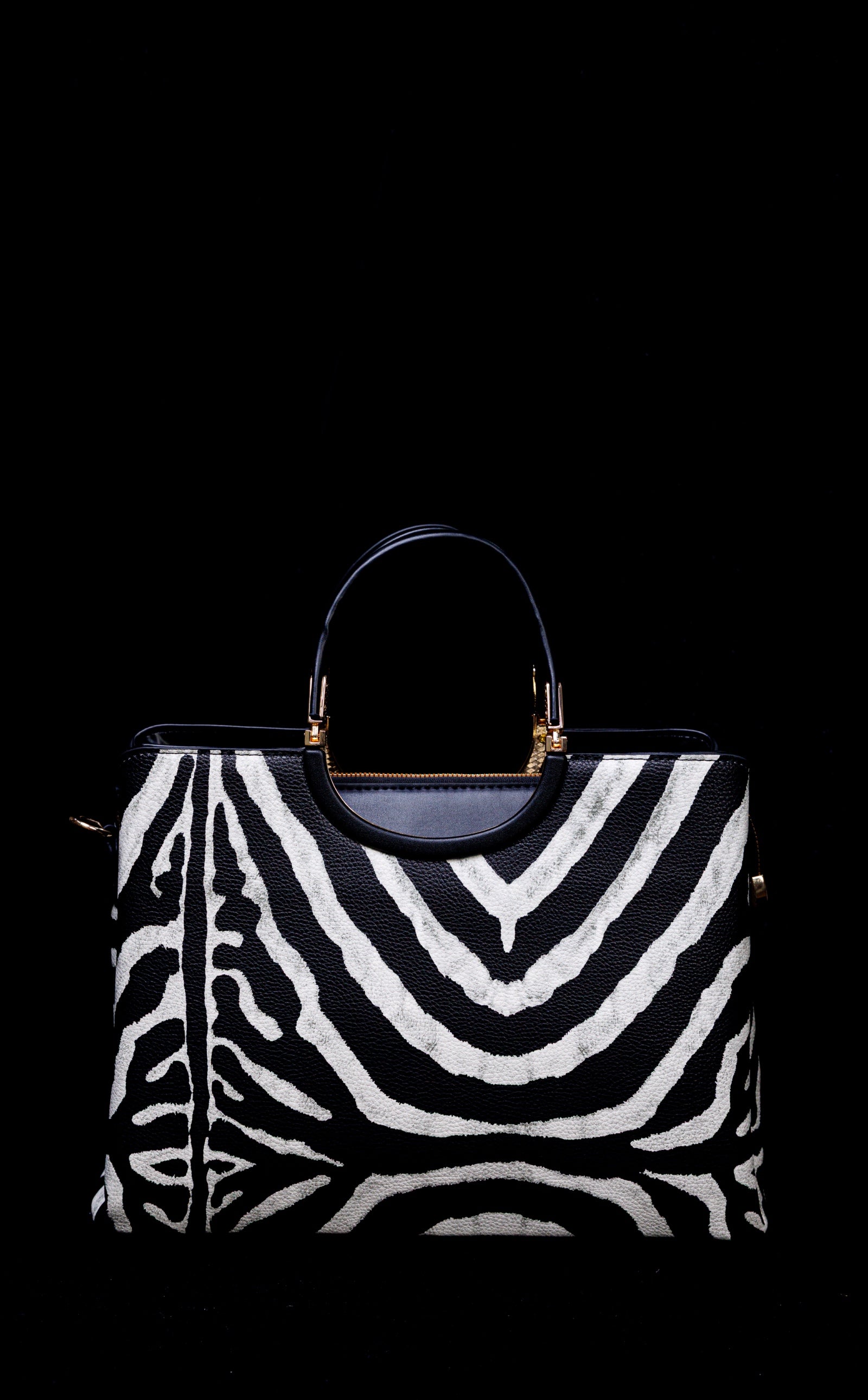 A structured handbag with a bold black and white zebra print pattern, featuring a dark navy blue leather top handle and trim with gold-tone hardware, set against a solid black background.