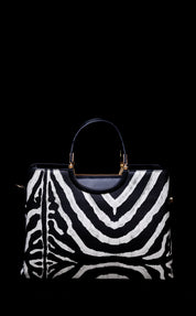 A structured handbag with a bold black and white zebra print pattern, featuring a dark navy blue leather top handle and trim with gold-tone hardware, set against a solid black background.