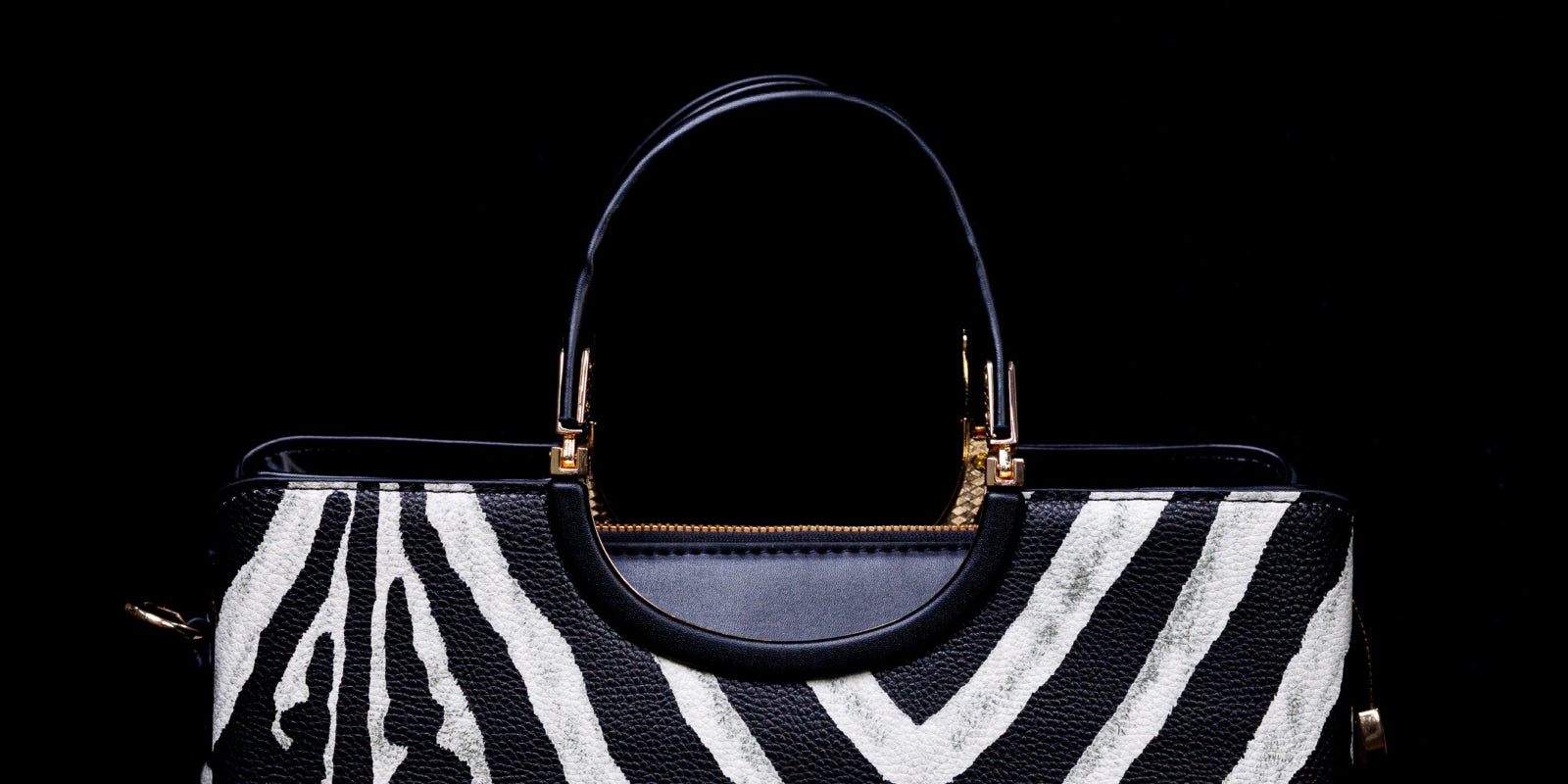 A structured handbag with a bold black and white zebra print pattern, featuring a dark navy blue leather top handle and trim with gold-tone hardware, set against a solid black background.
