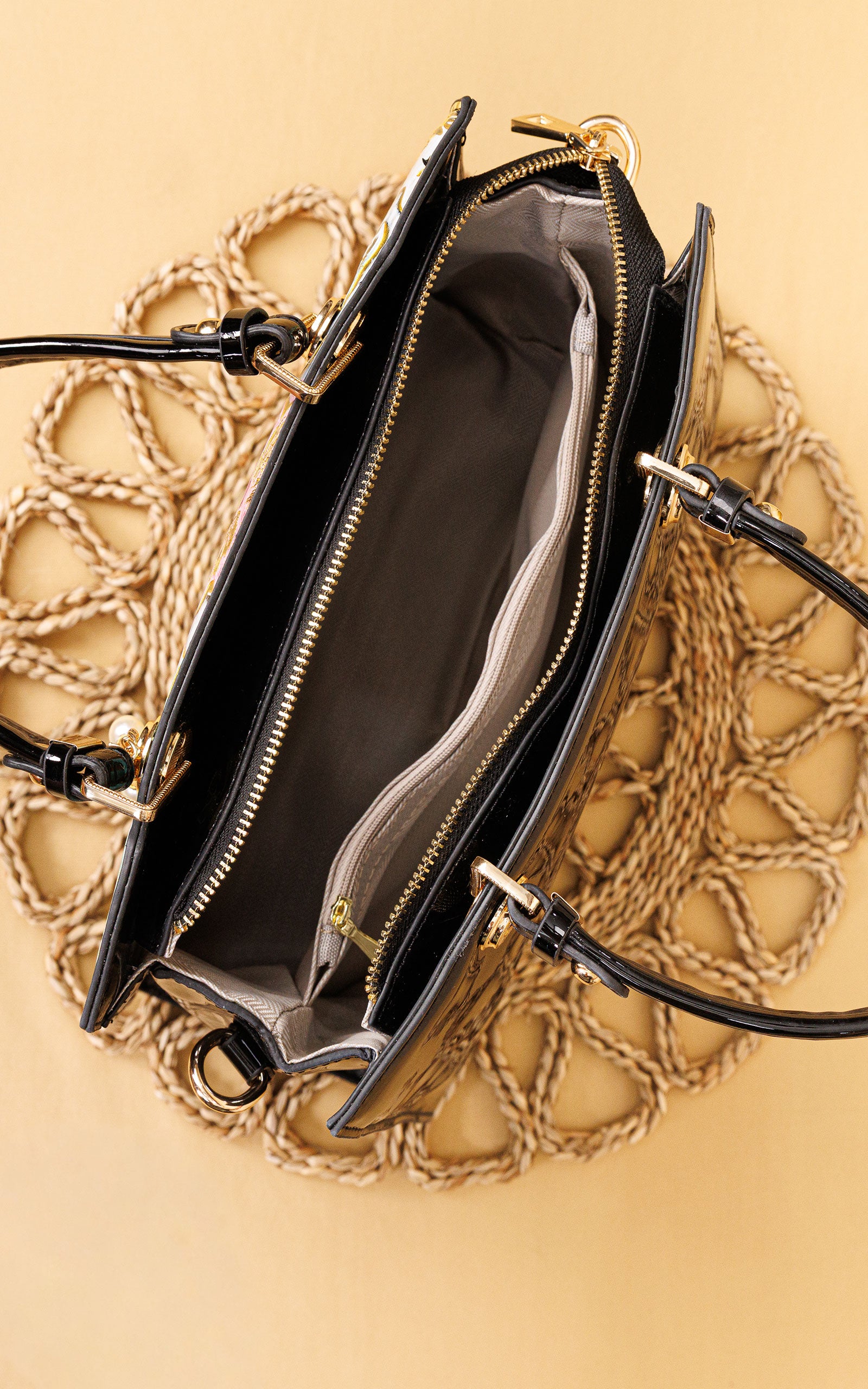 An open black leather handbag with gold-tone hardware, showing a dark gray interior and zippered compartments, laid flat on a beige woven placemat over a warm beige background.