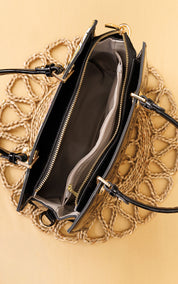 An open black leather handbag with gold-tone hardware, showing a dark gray interior and zippered compartments, laid flat on a beige woven placemat over a warm beige background.