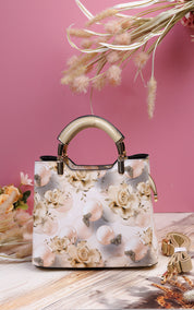 A structured handbag with a cream and beige floral print, featuring a curved gold-tone top handle and gold hardware, set against a pink background with decorative dried grass and a wooden surface.