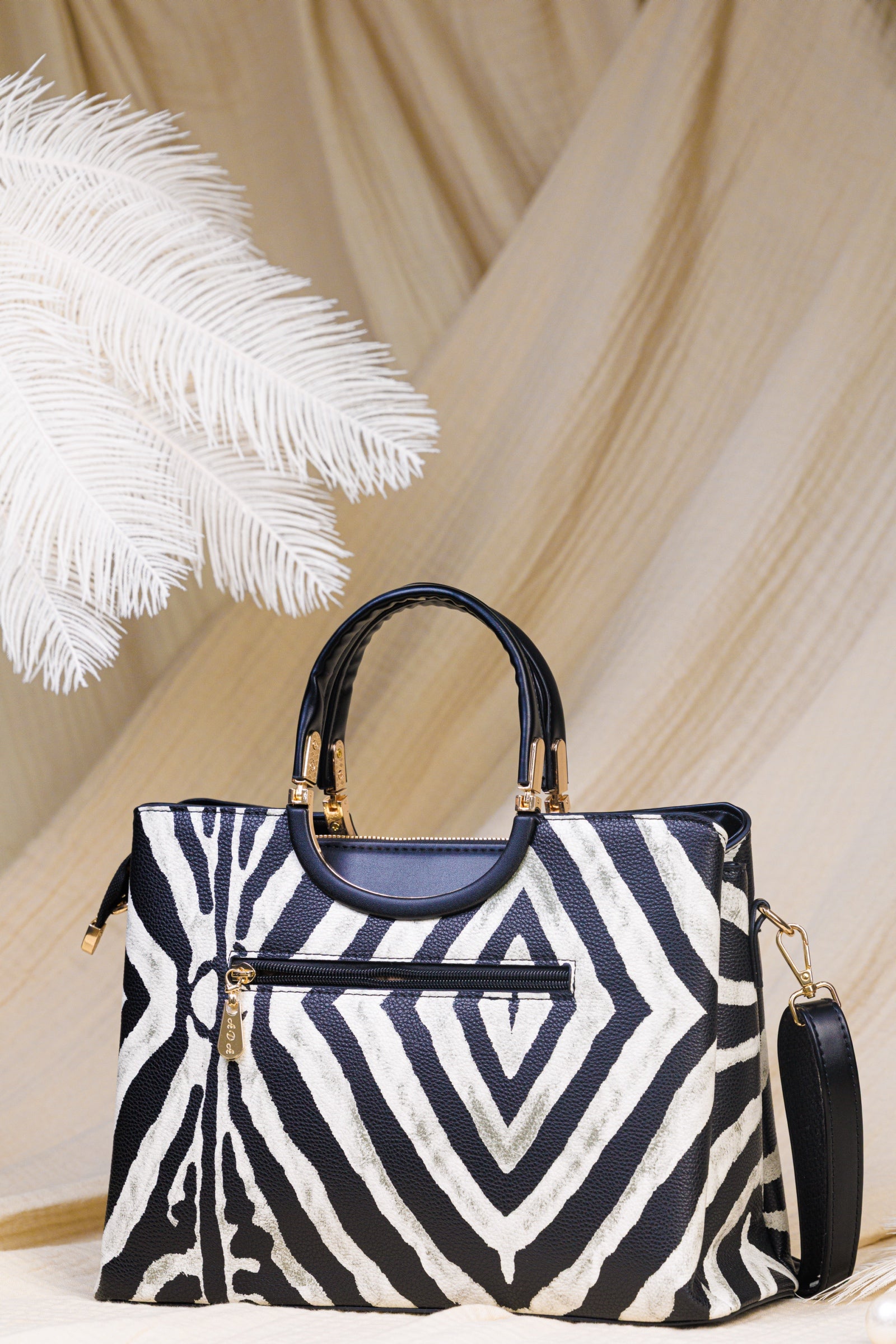 A structured handbag with a bold black and white geometric zebra pattern, featuring dark navy blue leather top handles and trim, gold-tone hardware, and a matching shoulder strap, set against a draped beige fabric backdrop with a white feather prop.