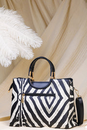 A structured handbag with a bold black and white geometric zebra pattern, featuring dark navy blue leather top handles and trim, gold-tone hardware, and a matching shoulder strap, set against a draped beige fabric backdrop with a white feather prop.
