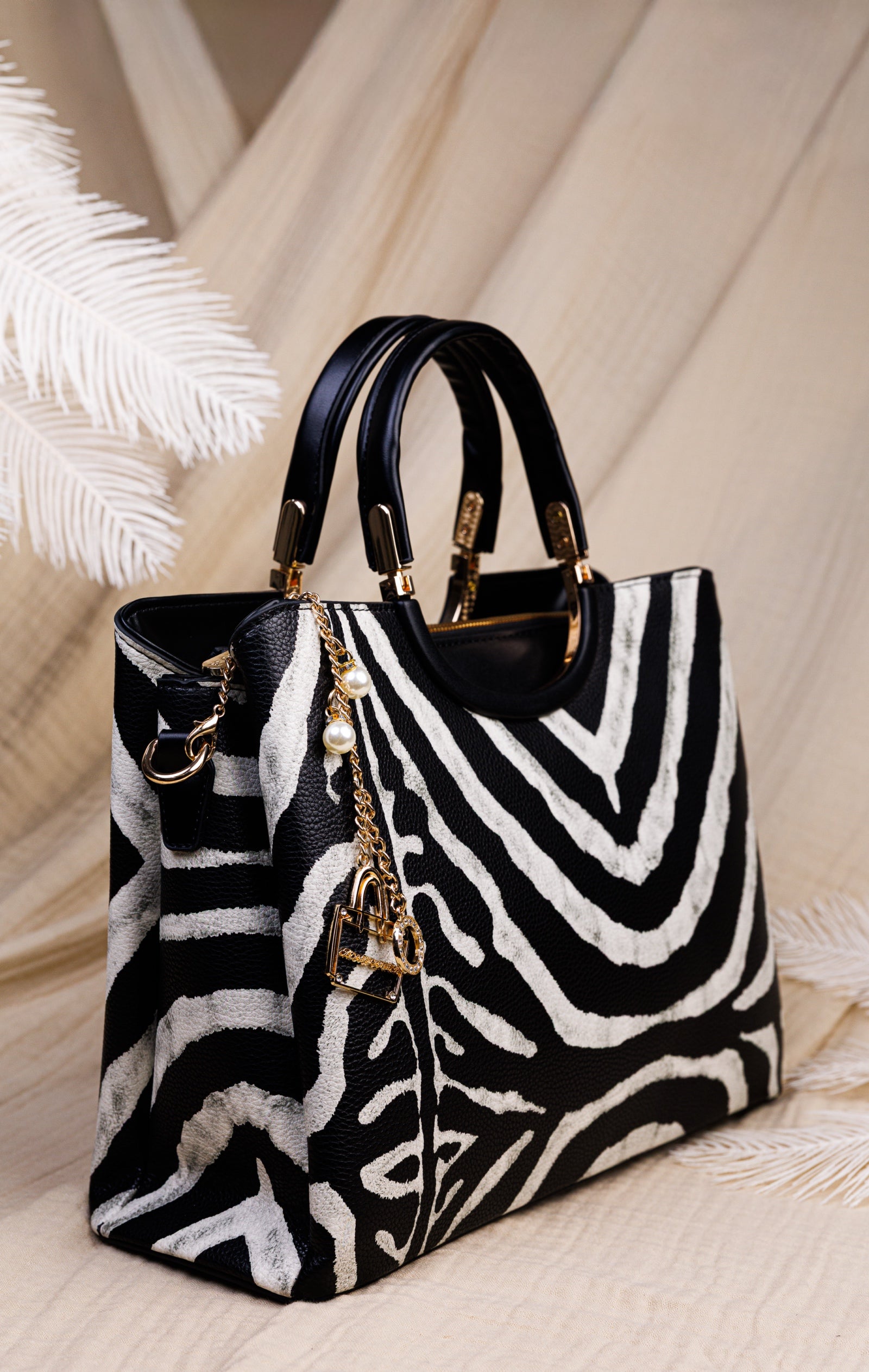A structured handbag with a black and white zebra print pattern, black leather handles, and gold-tone hardware, styled on a light beige fabric background with decorative white palm leaves.