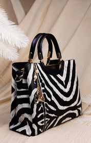 A structured handbag with a black and white zebra print pattern, black leather handles, and gold-tone hardware, styled on a light beige fabric background with decorative white palm leaves.